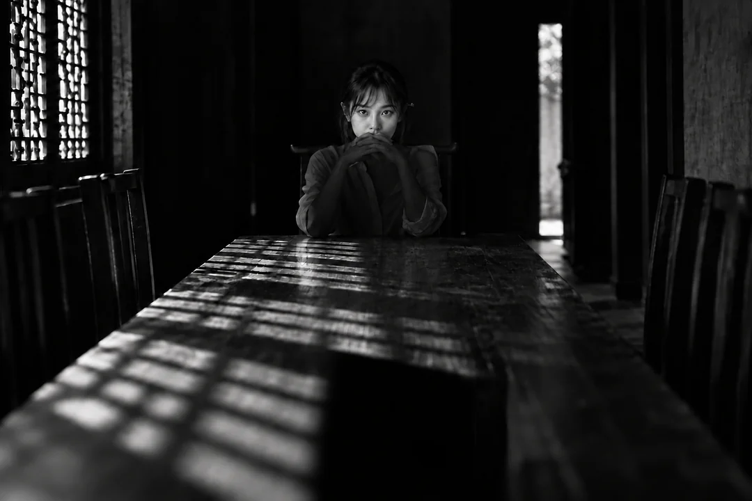 李瓶兒，黑白橫幅坐在長桌盡頭十指交握在唇前只有眼睛受光格柵光橫過桌面白襯衫（AI 生成，非真人照片）